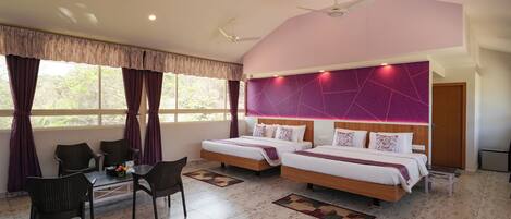 A bedroom with two beds, a dining area with chairs, a ceiling fan, and a view of trees through the window.