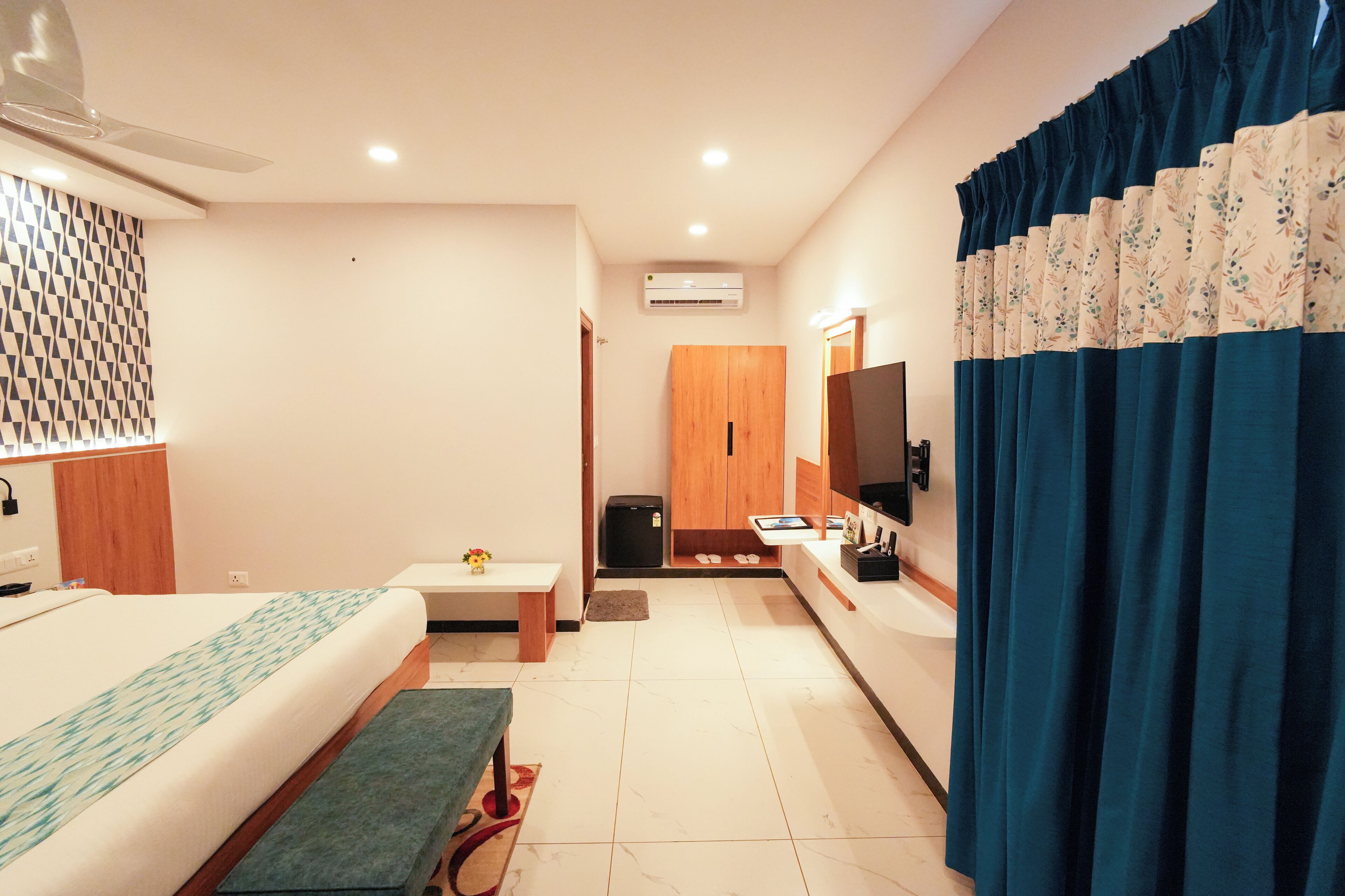 Habitación de hotel con cama, una mesita, un banco, televisor y baño visible a través de una puerta abierta.