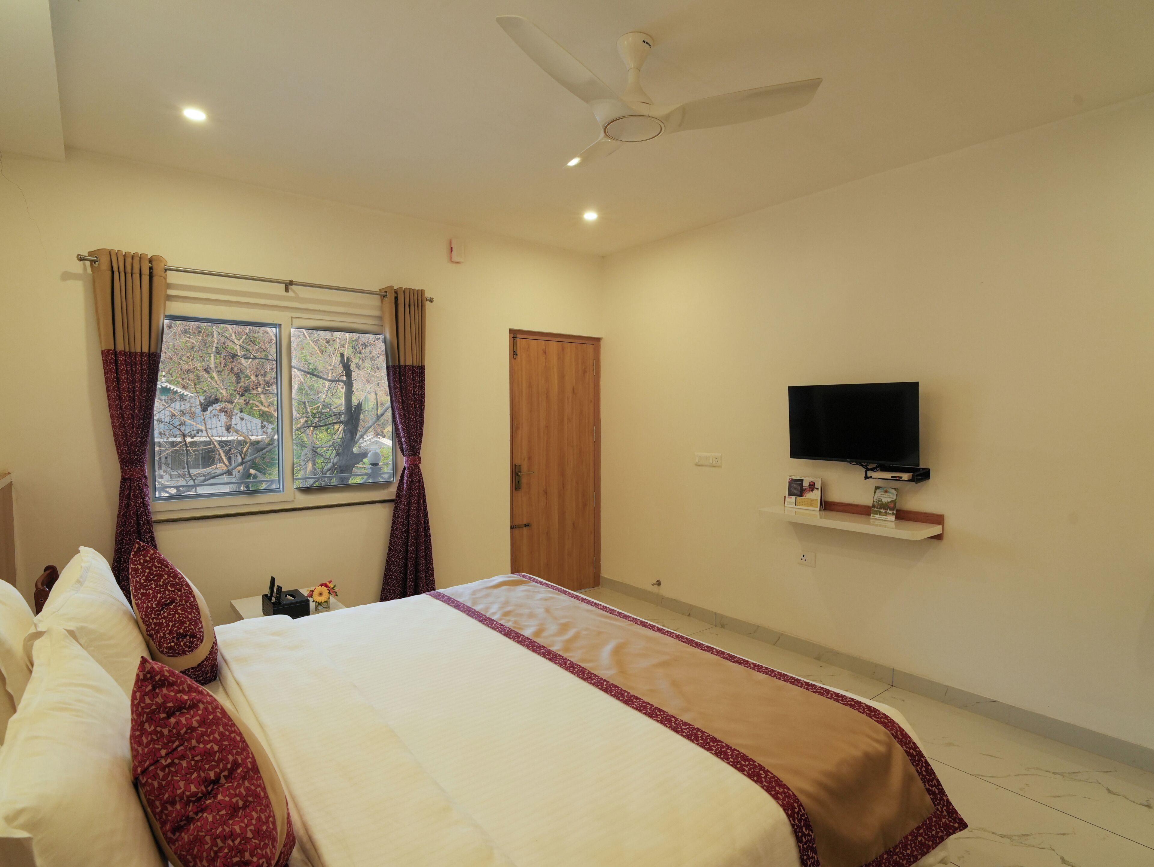 A hotel room with a bed, a ceiling fan, a TV mounted on the wall, and a window with curtains.