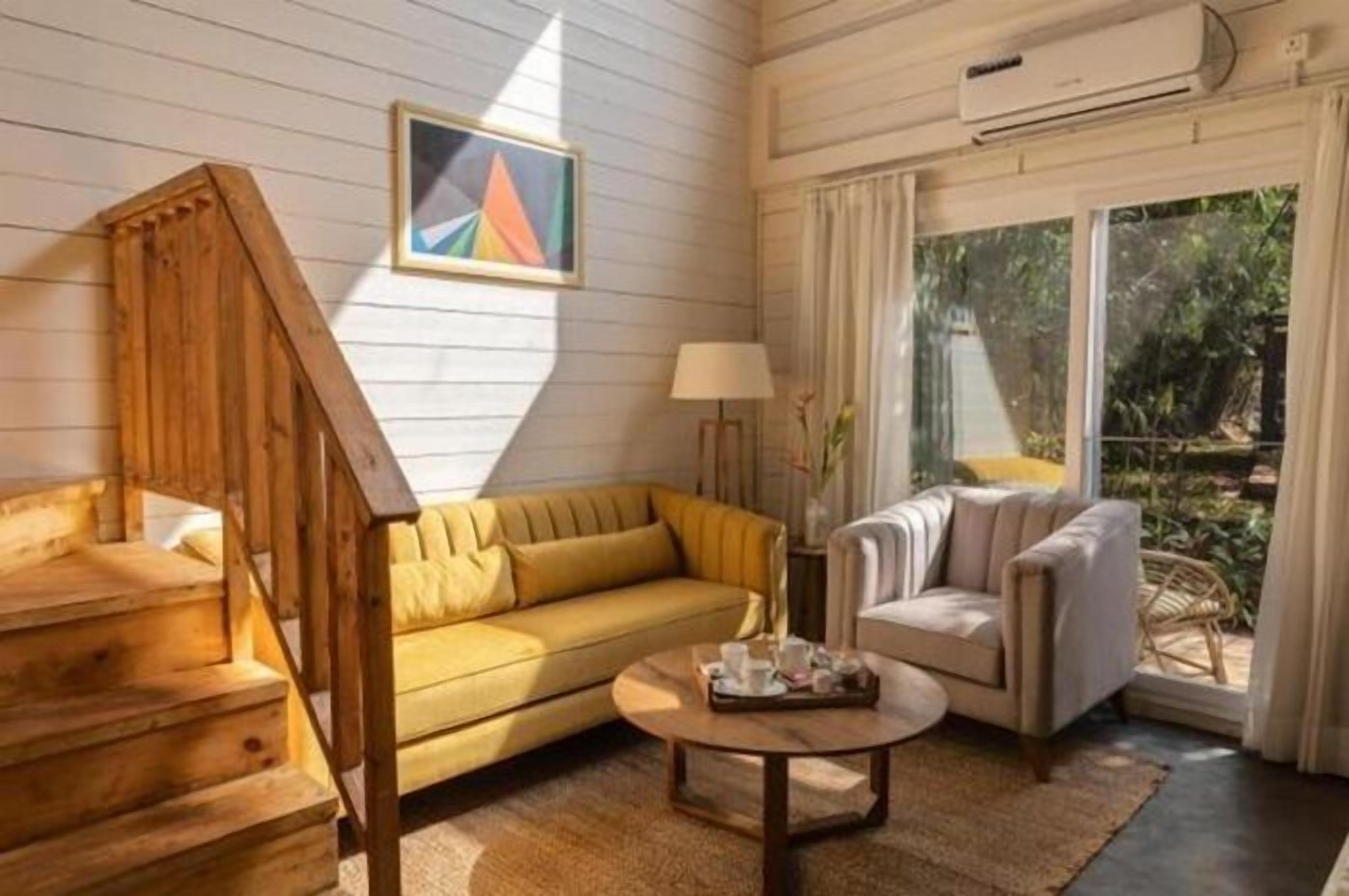 A room with a wooden staircase, a yellow sofa, a grey armchair, a round coffee table, and a large window with curtains.