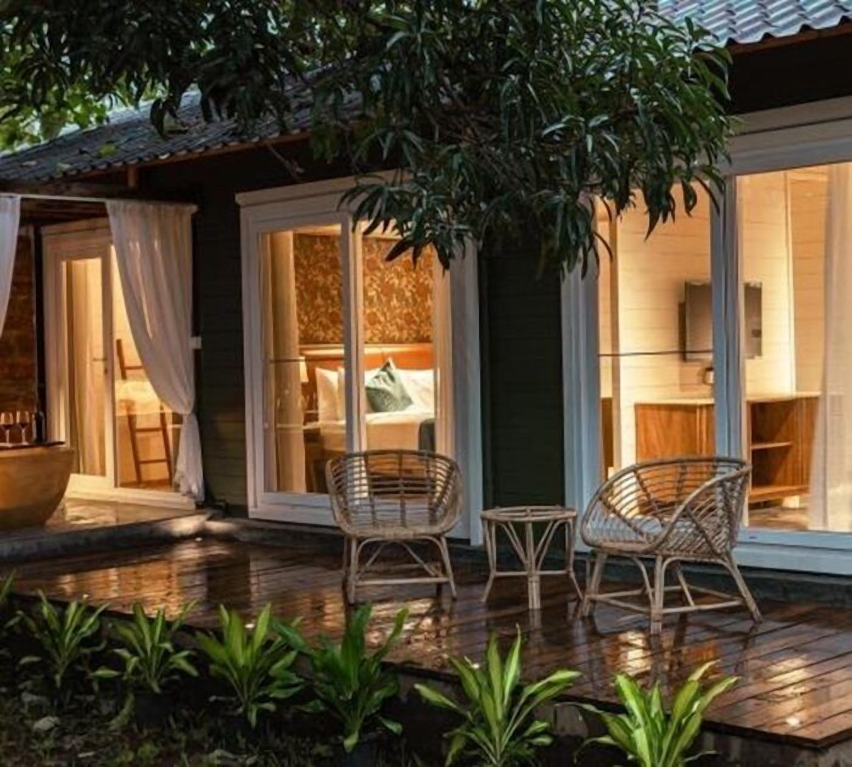 Une terrasse en bois avec des chaises en osier, une grande plante en pot et une vue sur l’intérieur à travers des portes vitrées.
