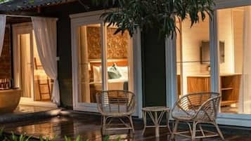 A wooden deck with wicker chairs, a large potted plant, and a view of the interior through glass doors.