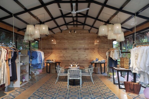 A rustic interior with wooden walls, a central table with chairs, hanging light fixtures, and clothing racks.