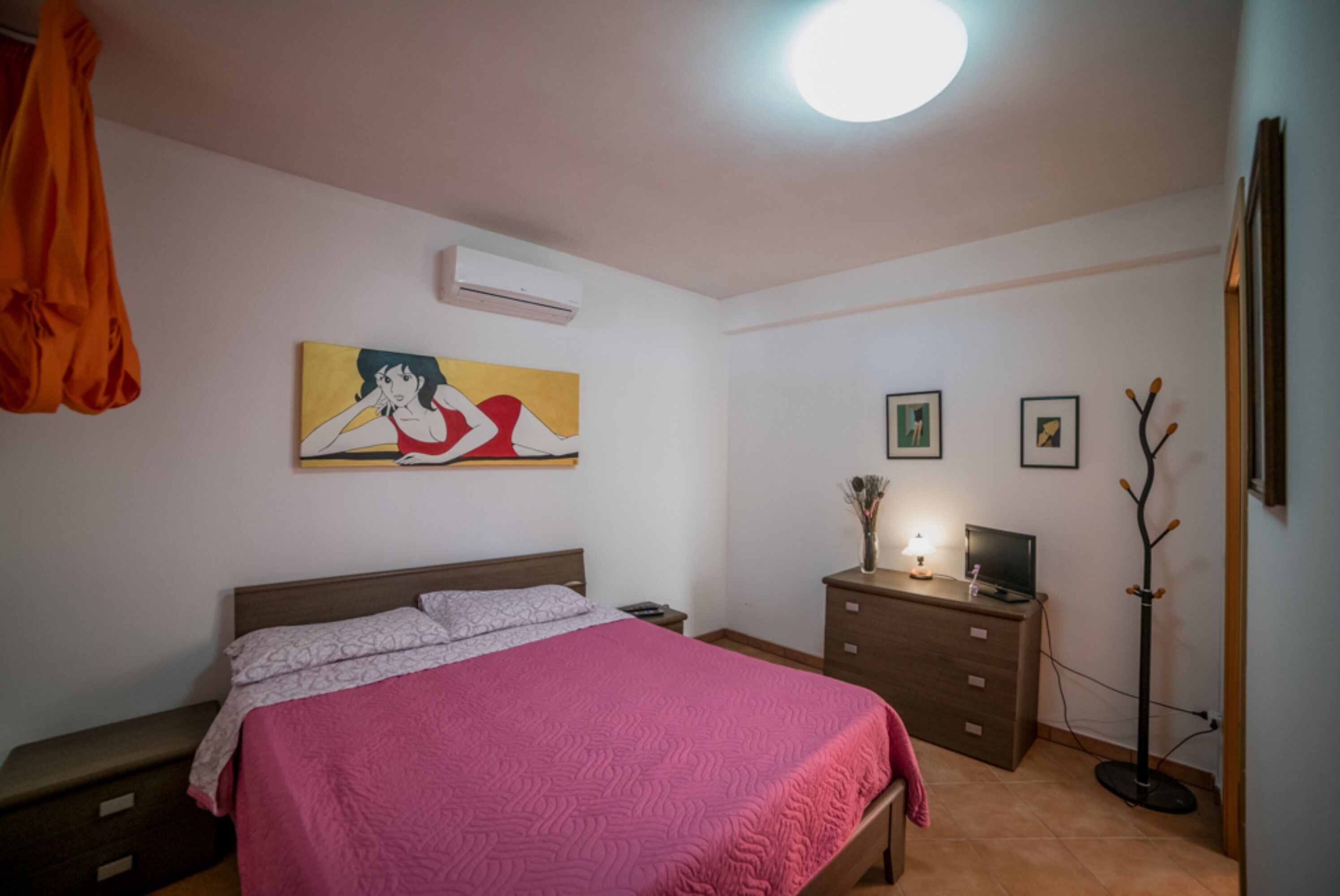 A bedroom with a bed, a dresser, an air conditioner, a lamp, and a coat rack.