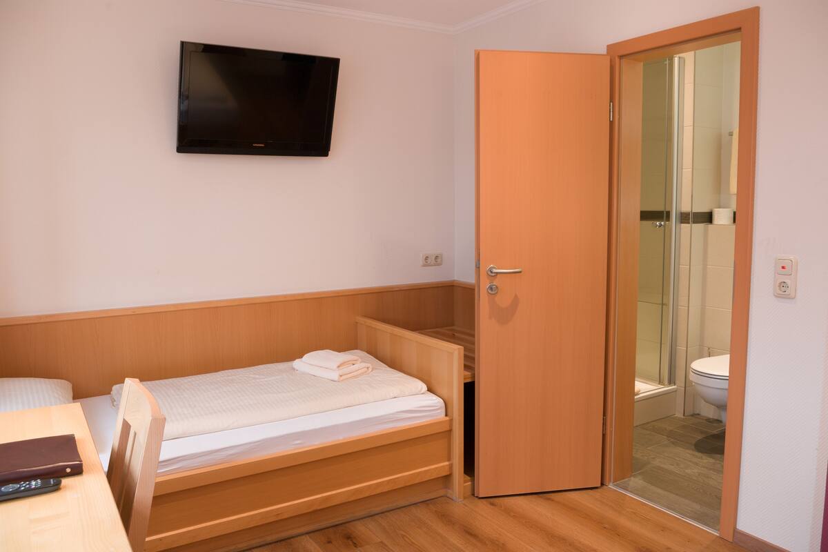 Una habitación pequeña con una cama individual, un televisor en la pared y una puerta que da a un baño.