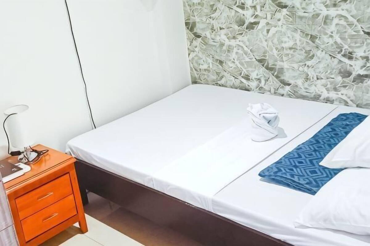 A single bed with a white mattress and a blue patterned blanket, a wooden nightstand with a lamp, and a wall with a textured design.