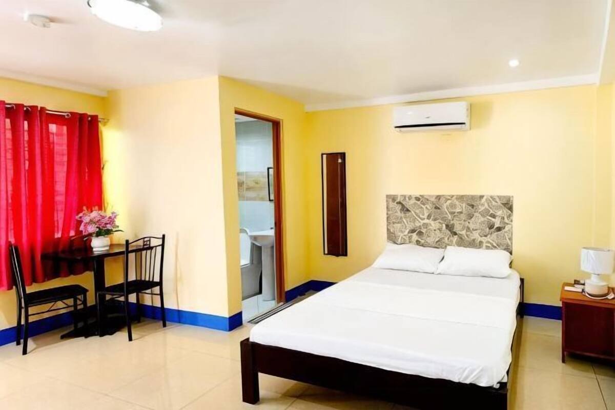 A hotel room with a bed, a desk with a chair, a small table with a vase, a nightstand, and an air conditioning unit.