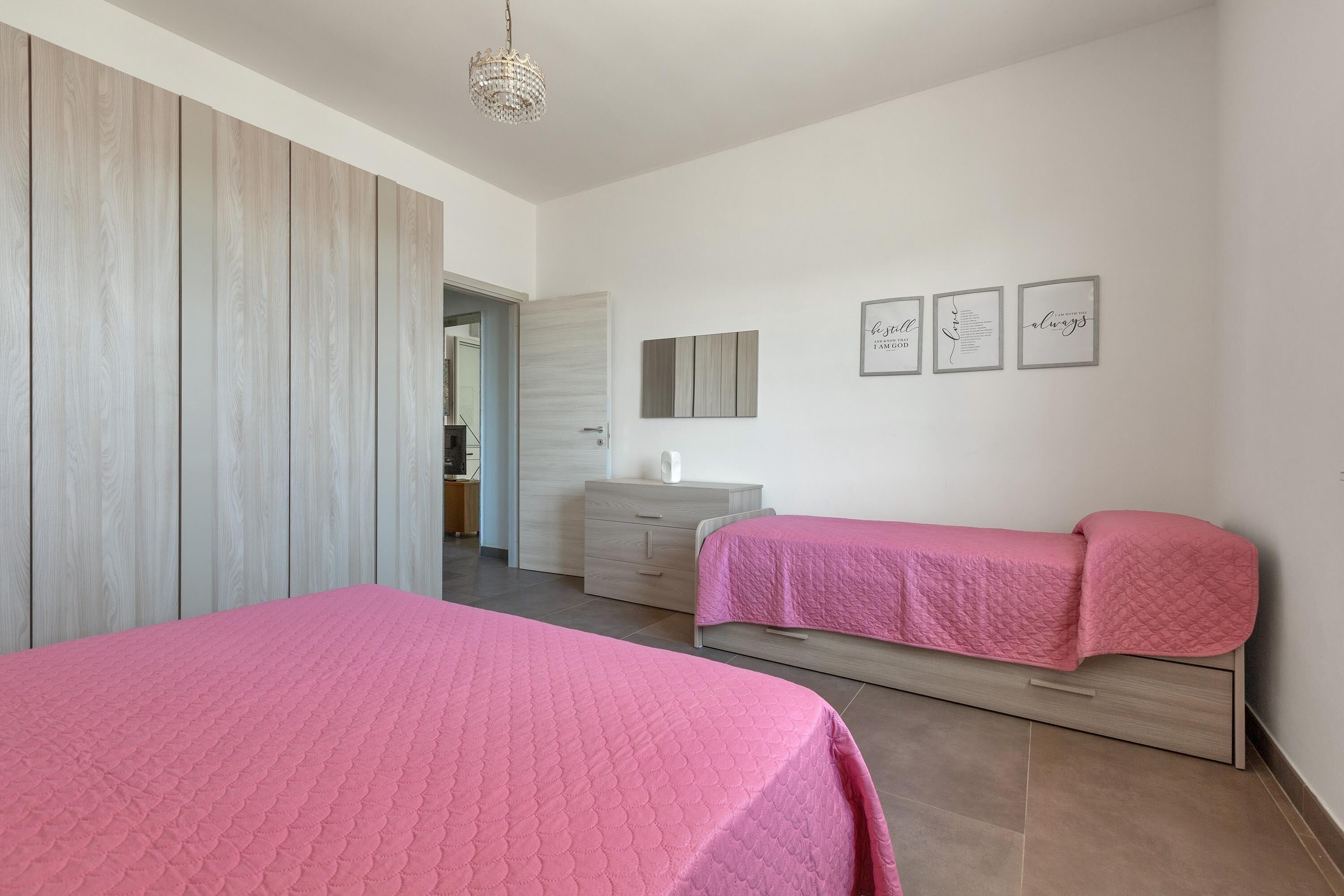 A bedroom with two beds, a wooden wardrobe, a bedside table, and a wall with framed artwork.