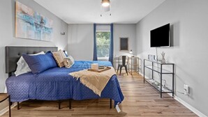 Ein Schlafzimmer mit Bett, Deckenventilator, Fernseher, Schreibtisch und einem Fenster mit Vorhängen.