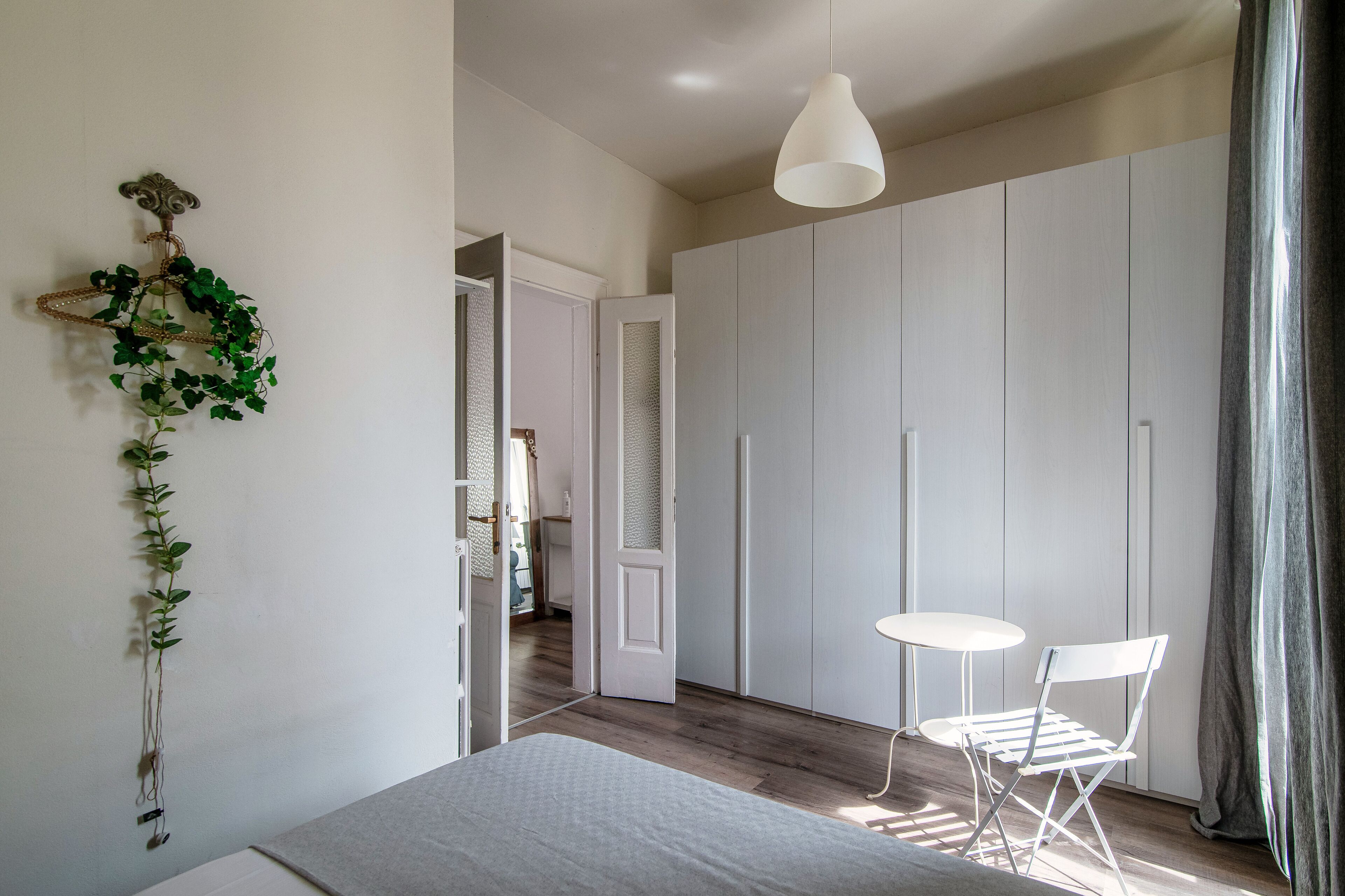 Un dormitorio con cama, una silla blanca, una mesita, una planta colgante y un armario.