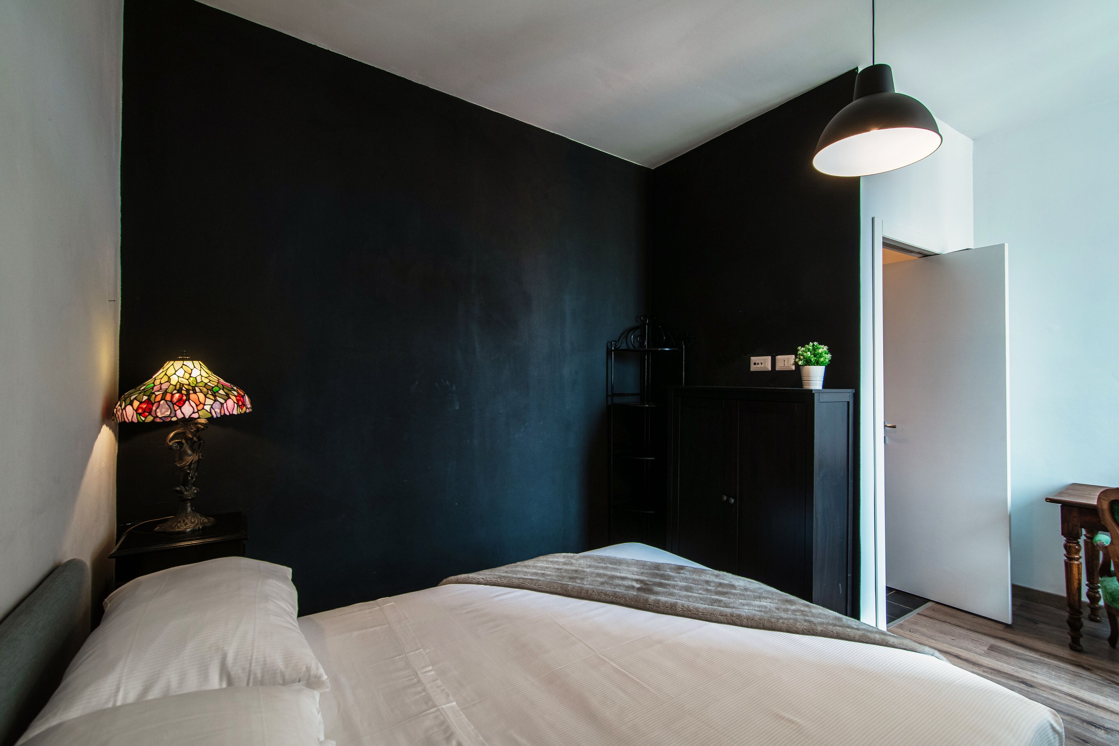 Quarto com cama, mesa de cabeceira com abajur, guarda-roupa e porta para outro cômodo.