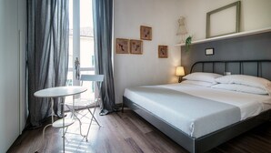 A neatly arranged hotel room with a bed, a small table, and a chair. There are curtains, a lamp, and framed pictures on the wall. - Glamour (Como)