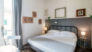 A neatly made bed with white linens, a bedside table with a lamp, and a window with curtains. - Glamour (Como)