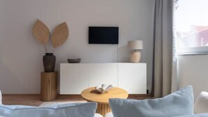 Een moderne kamer met een witte bank, een ronde houten tafel, een wit kastje en een flatscreen televisie aan de muur.