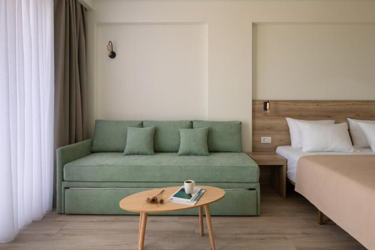 A room with a green sofa, a wooden nightstand, a bed with white bedding, and a small round table with a book and a cup.
