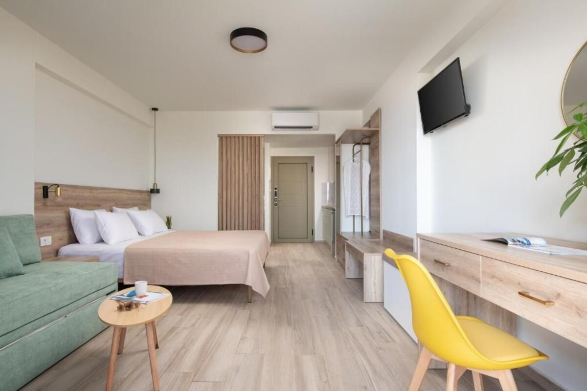 A modern hotel room with a bed, a desk, a chair, a small table, a TV, and a plant.