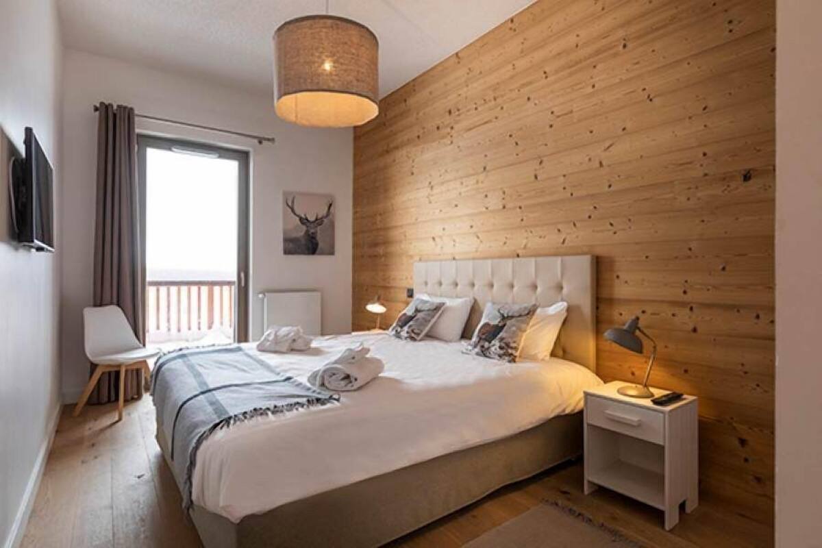 Et moderne soveværelse med en stor seng, træpaneler, en loftlampe, et natbord med lampe og et vindue med gardiner.