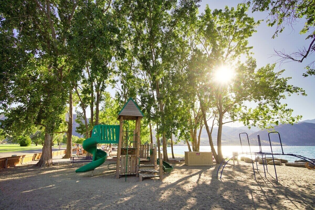 A playground with a slide, climbing structures, and a small shelter, surrounded by trees and a lake view.