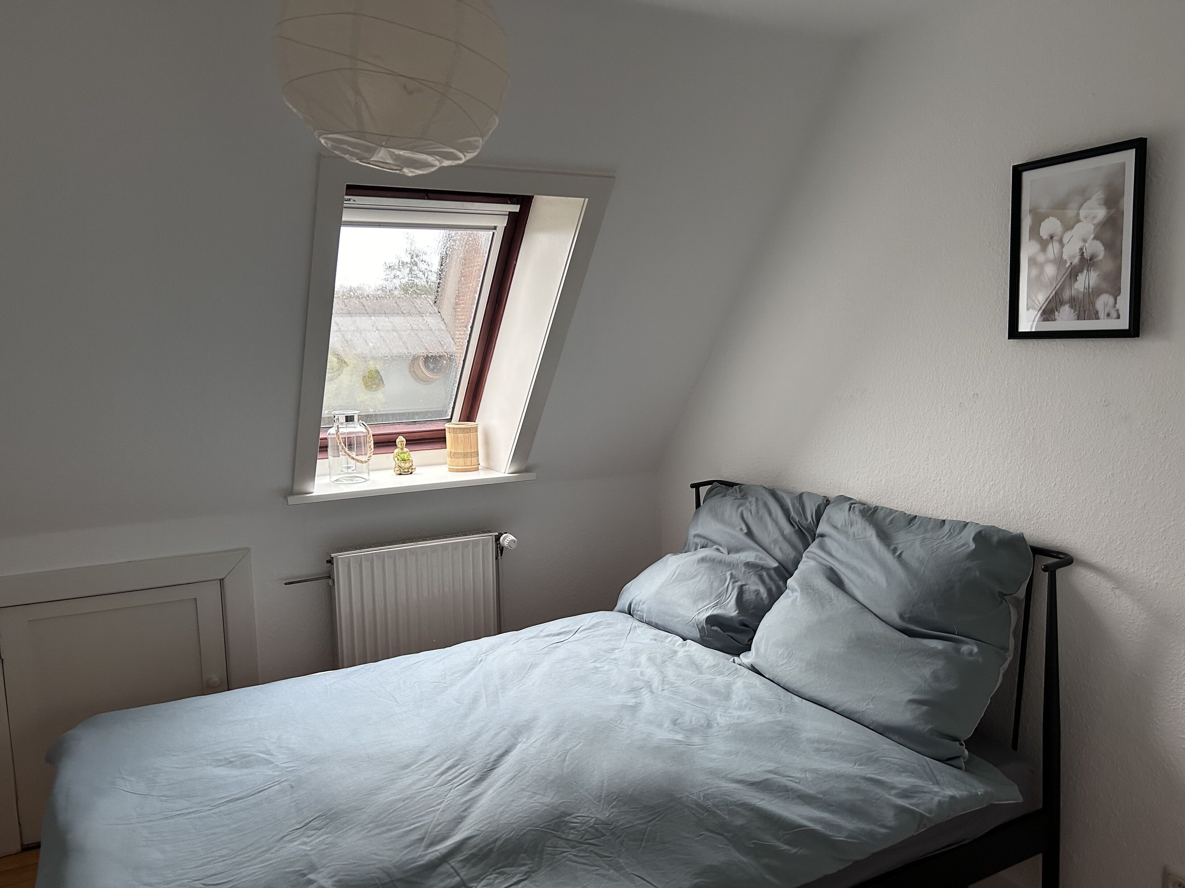 Un dormitorio con cama, ventana, radiador y un cuadro en la pared.