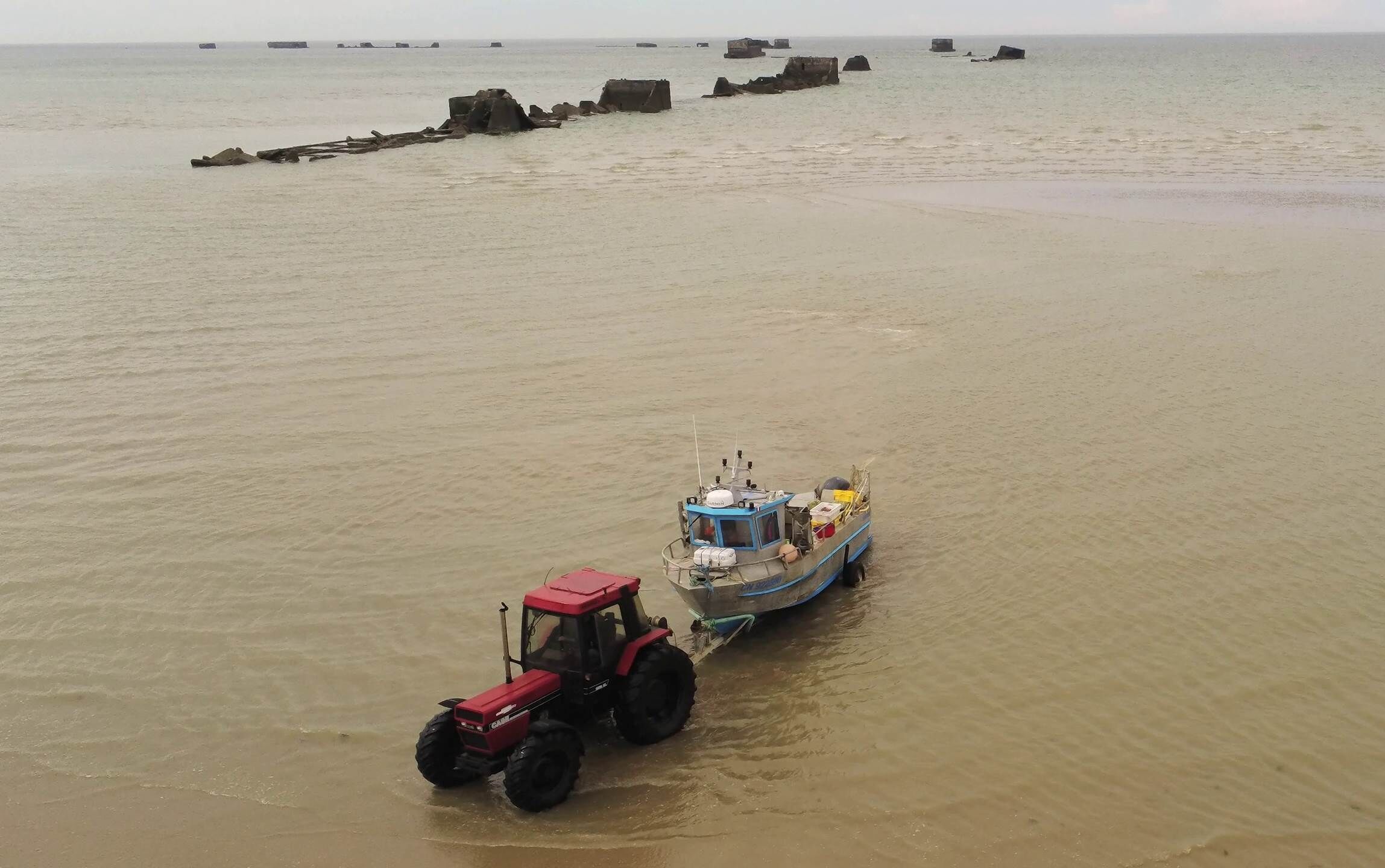 A red tractor towing a blue and yellow boat through a shallow water body with a sandy shore and scattered rocks.