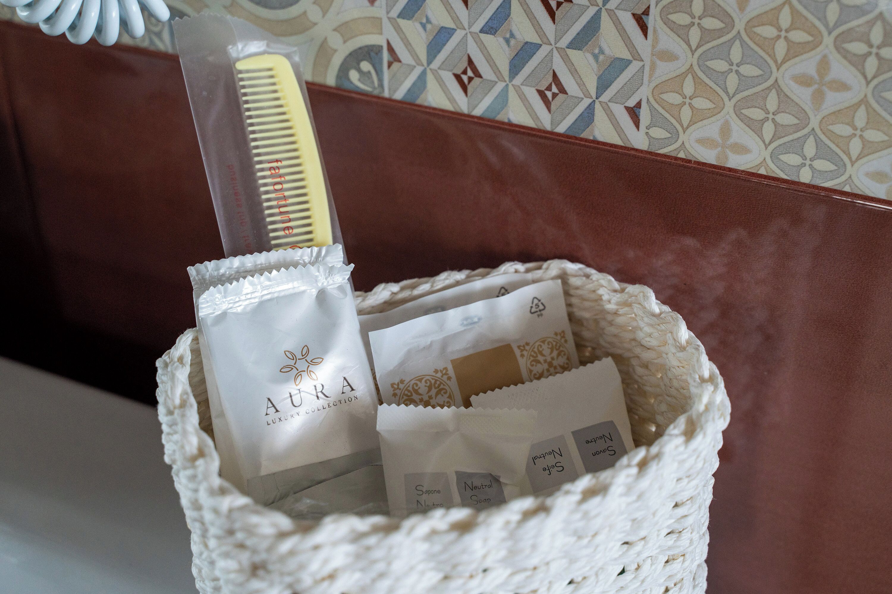 A woven basket containing a white bag with 'AURA LUXURY COLLECTION' branding, a comb, and several smaller white bags with product labels.