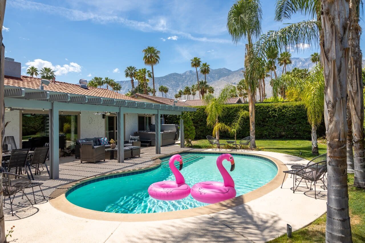 A swimming pool with two pink flamingo floats, surrounded by palm trees and a covered patio area with outdoor furniture.