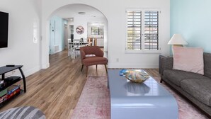 A modern living room with a flat-screen TV, a sofa, a coffee table, and a dining area in the background. - Palm Springs Oasis: Gated Condo w/ Pool & Hot Tub (Palm Springs)