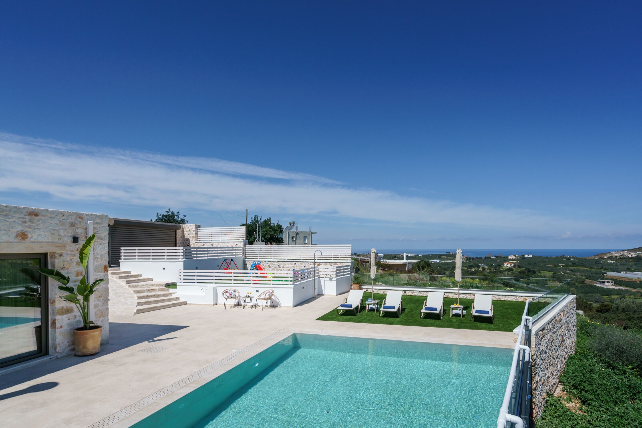 A modern outdoor pool area with a clear view of the sea, a stone wall, and a seating area with a table and chairs.