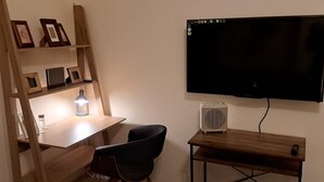 Ein Zimmer mit einem Holztisch, einem schwarzen Stuhl, einem an der Wand befestigten Fernseher und einem kleinen weißen Ventilator auf einem Tisch.
