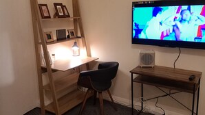 Ein kleiner, gut beleuchteter Raum mit einem Holztisch, einem Stuhl, einem an der Wand montierten Fernseher und einem Regal mit gerahmten Bildern.