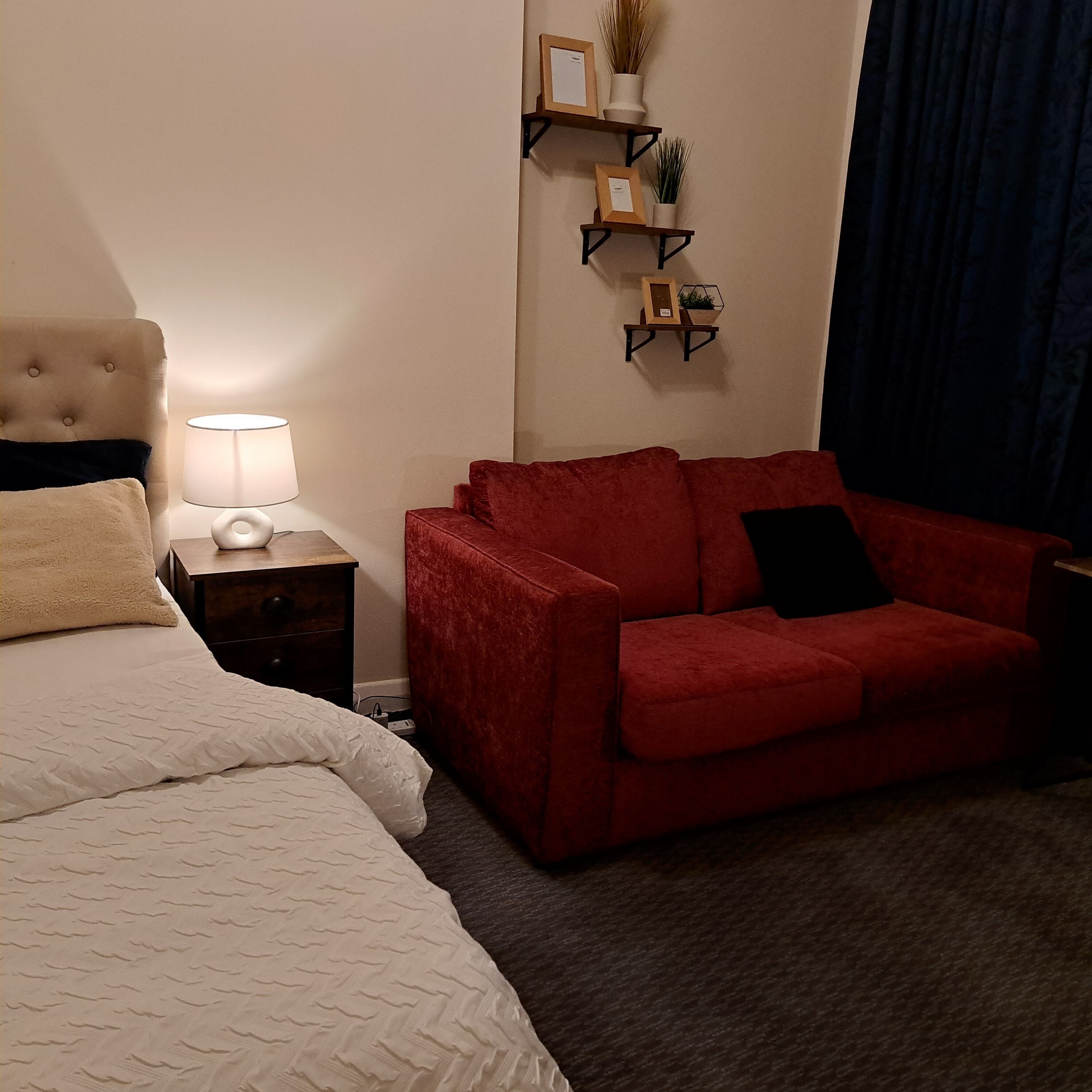 A hotel room with a bed, a red sofa, a nightstand with a lamp, and wall-mounted shelves with decorative items.