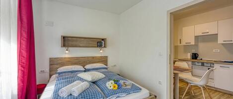 Un dormitorio con cama, mesita de noche, cabeza de cama de madera, ventana con cortinas y vista a un área de cocina.