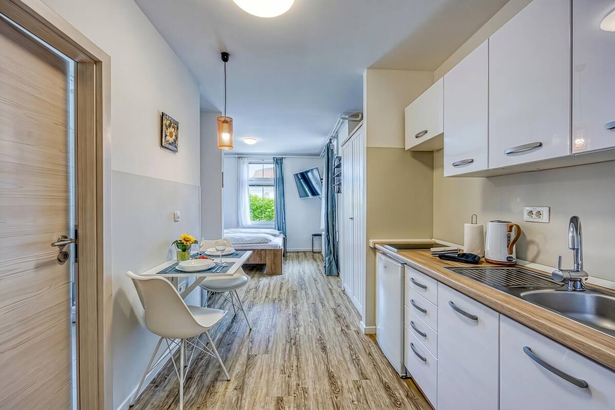 Un estrecho pasillo que conduce a un dormitorio con una cama, una pequeña mesa con sillas, un ventanal con cortinas y una kitchenette con un lavabo y armarios.