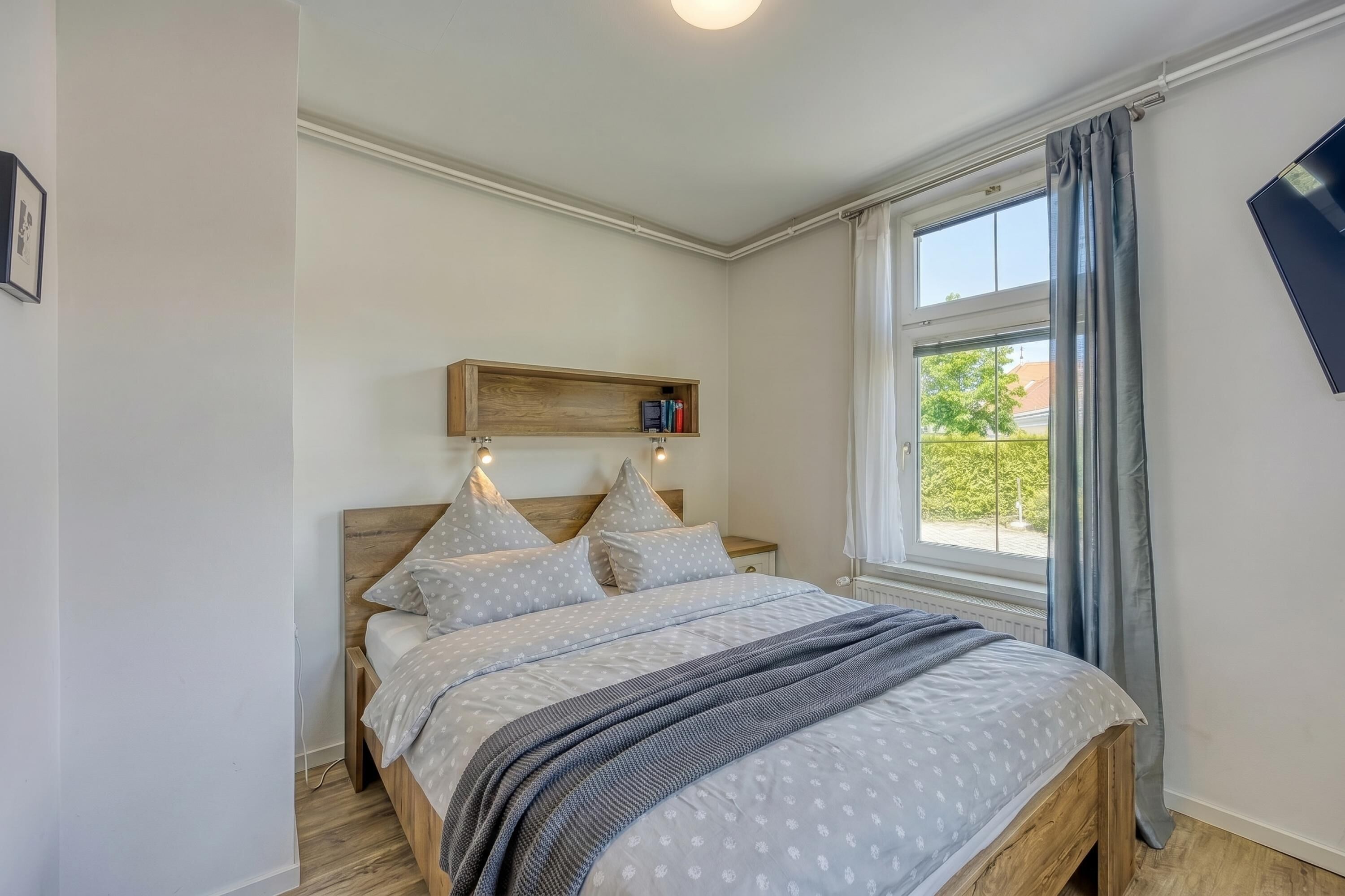 Un dormitorio con cama, mesita de noche, estante sobre la cama y ventana con cortinas.