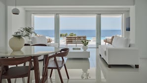 A modern living room with a glass wall overlooking the sea, a dining area with a glass table, and a white sofa. - OLYN VILLAS (Nikiti)