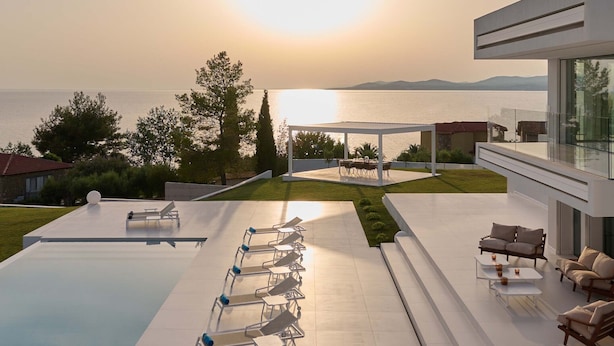 A modern outdoor pool area with lounge chairs and a dining setup, overlooking a body of water and hills. - OLYN VILLAS (Nikiti)