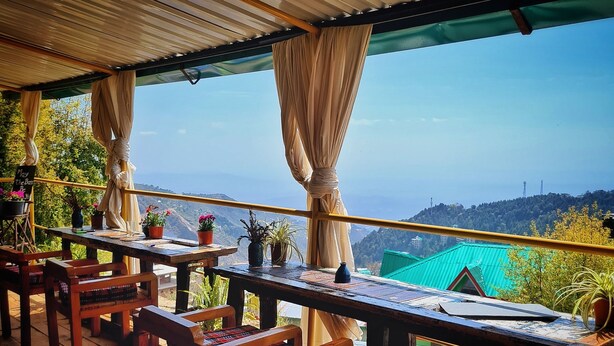 A wooden deck with a table and chairs, overlooking a scenic landscape with hills and a clear sky. - EDM Hostel (Dharamshala)