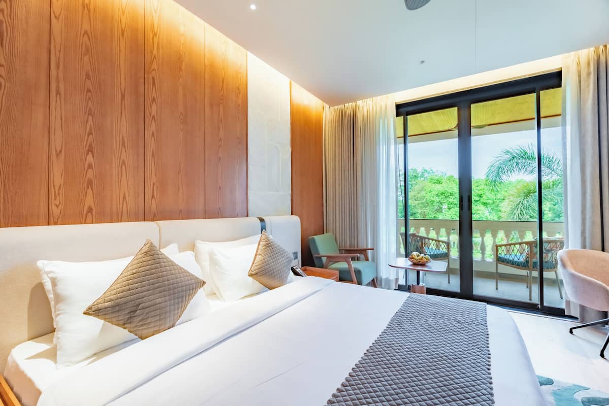 Une chambre d’hôtel avec un grand lit, un balcon doté d’une table et de chaises, et une vue sur la verdure.