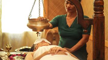 A person receiving a traditional massage with a wooden massage table and a brass oil dispenser.