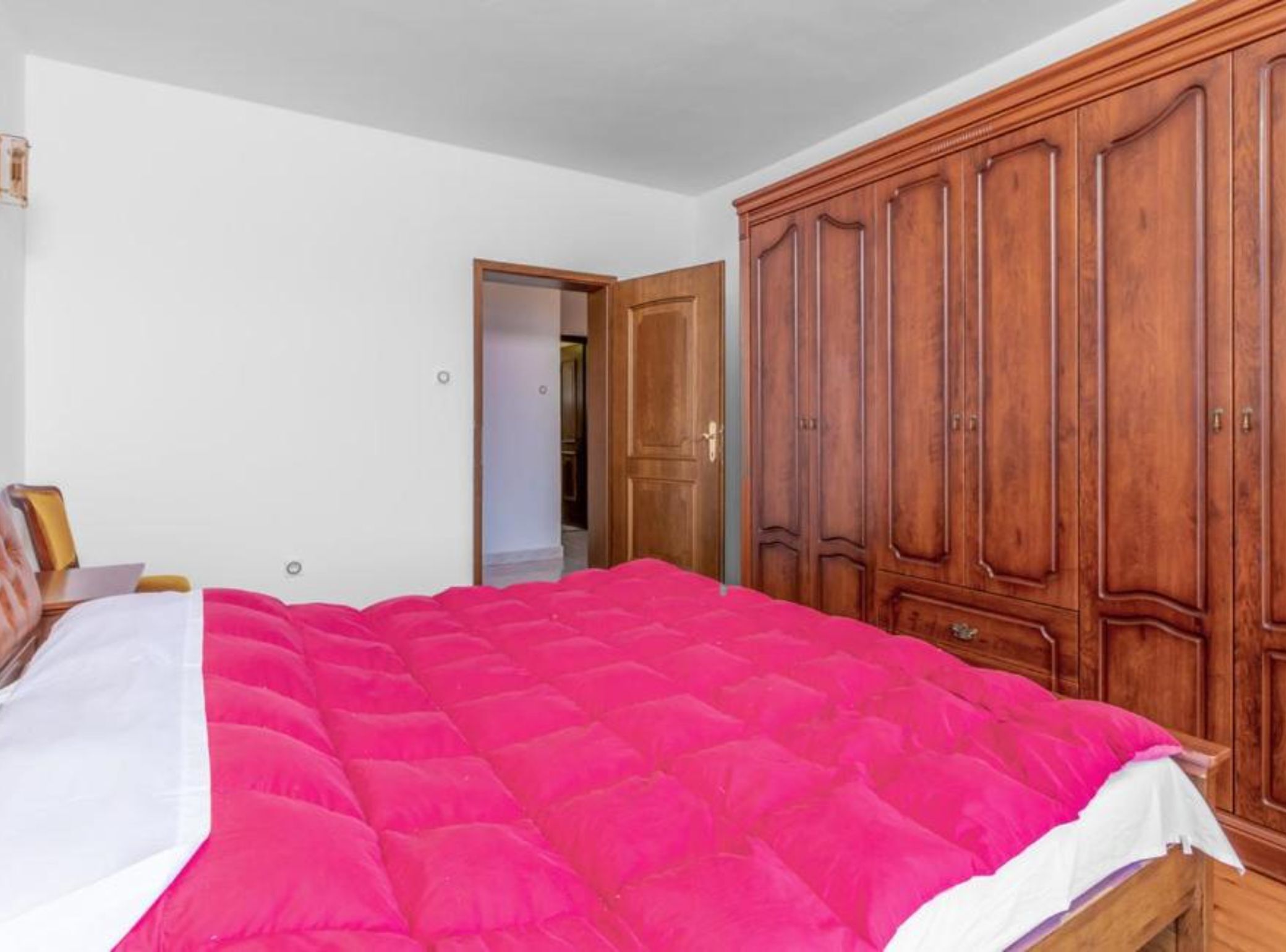 Un dormitorio con un armario grande de madera, una cama con colcha rosa y una pared blanca.