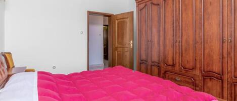 Un dormitorio con un armario grande de madera, una cama con colcha rosa y una pared blanca.