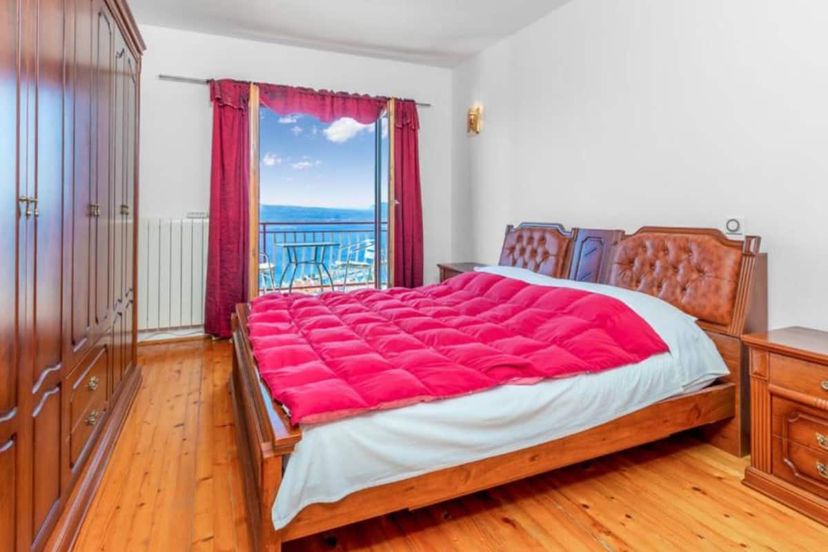 A bedroom with a wooden bed, a wooden wardrobe, and a view of the sea through a window with red curtains.