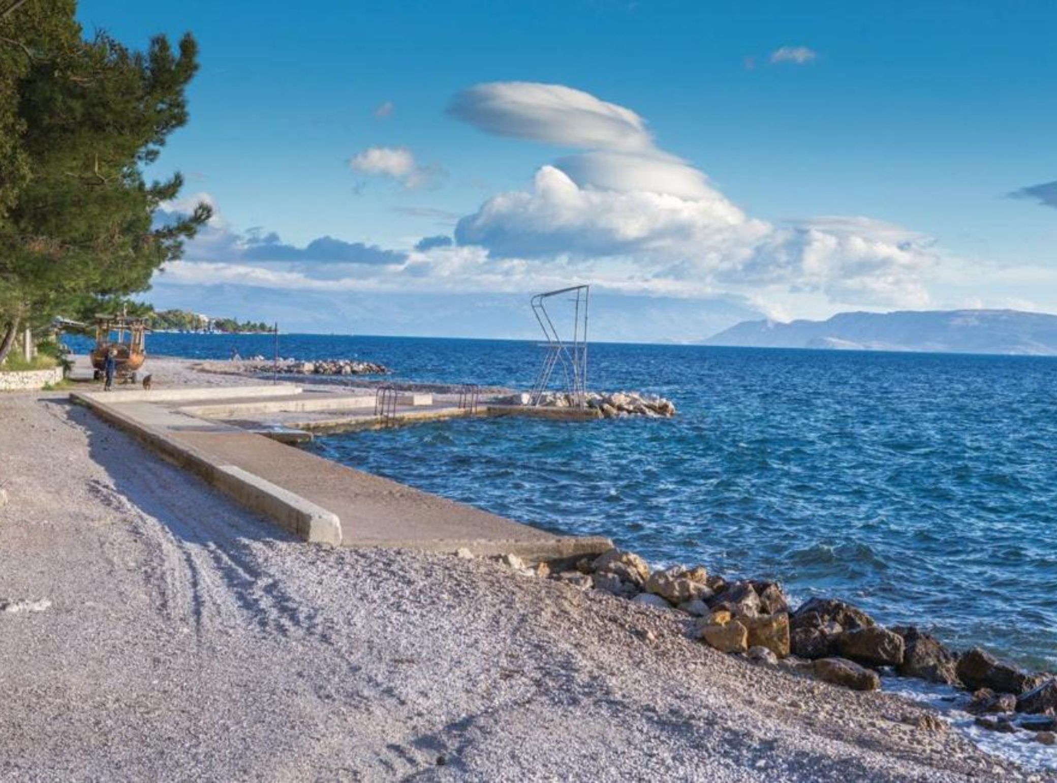A coastal road with a rocky shoreline, a pier extending into the sea, and a clear sky.