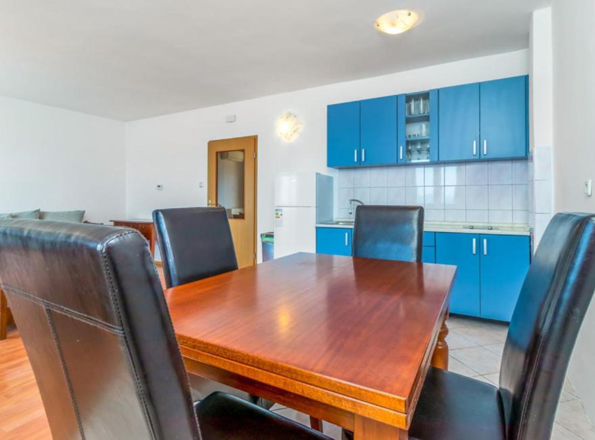 A dining area with a wooden table and black leather chairs, a kitchen with blue cabinets, and a white tiled backsplash.