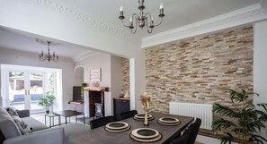 A dining area with a modern table set for four, a chandelier, and a brick-patterned wall. - Modern Interior | 5 Mins to Metro Centre | WiFi (Newcastle Upon Tyne)
