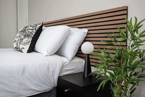 A bed with white bedding, a black and white pillow, a round white object on a black stand, and a green plant beside the bed.