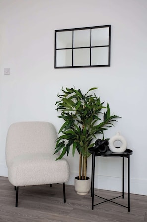 A room with a white chair, a potted plant, a small table with a white vase, and a framed picture on the wall.