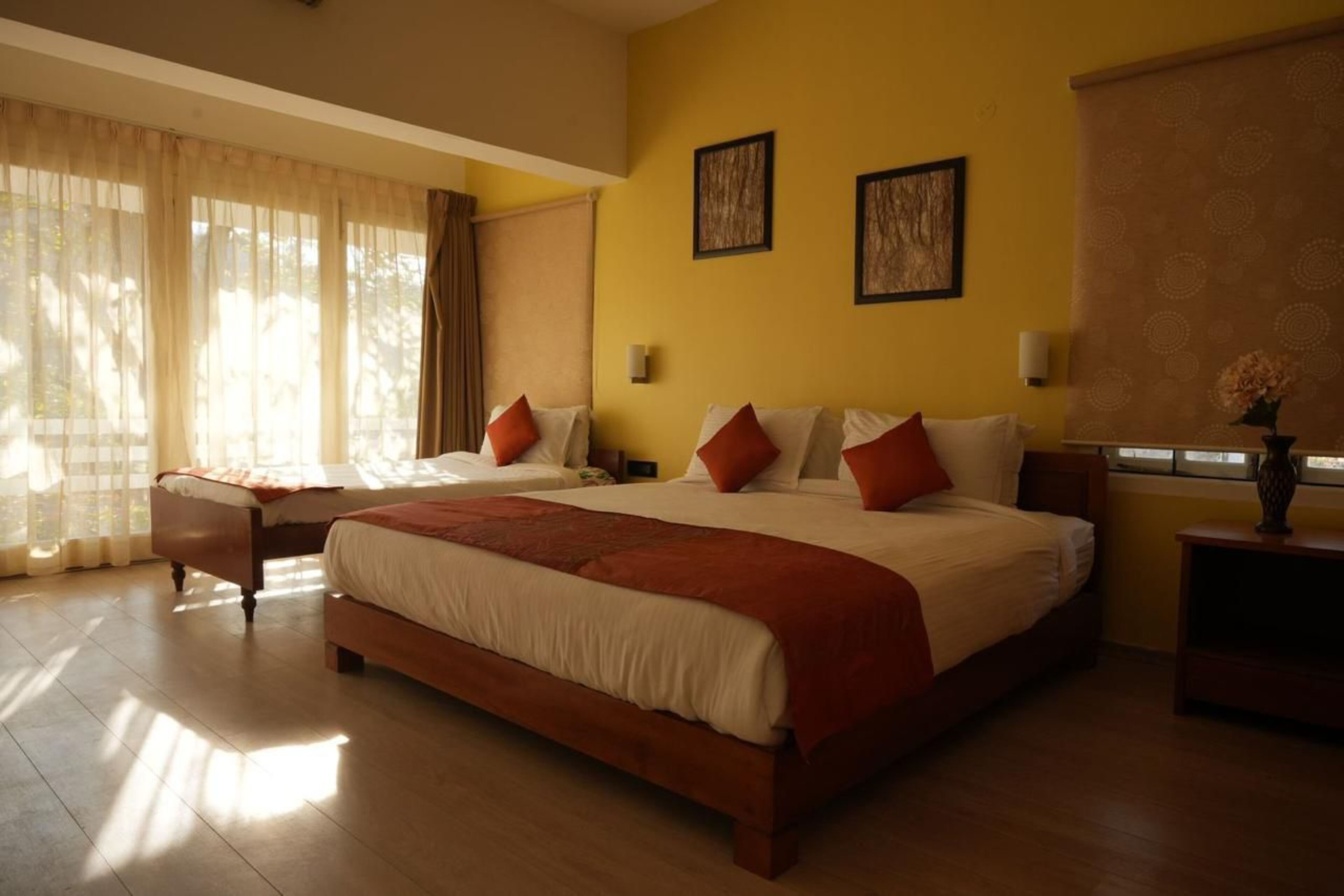 A hotel room with two beds, a large window with sheer curtains, and a small table with a vase.