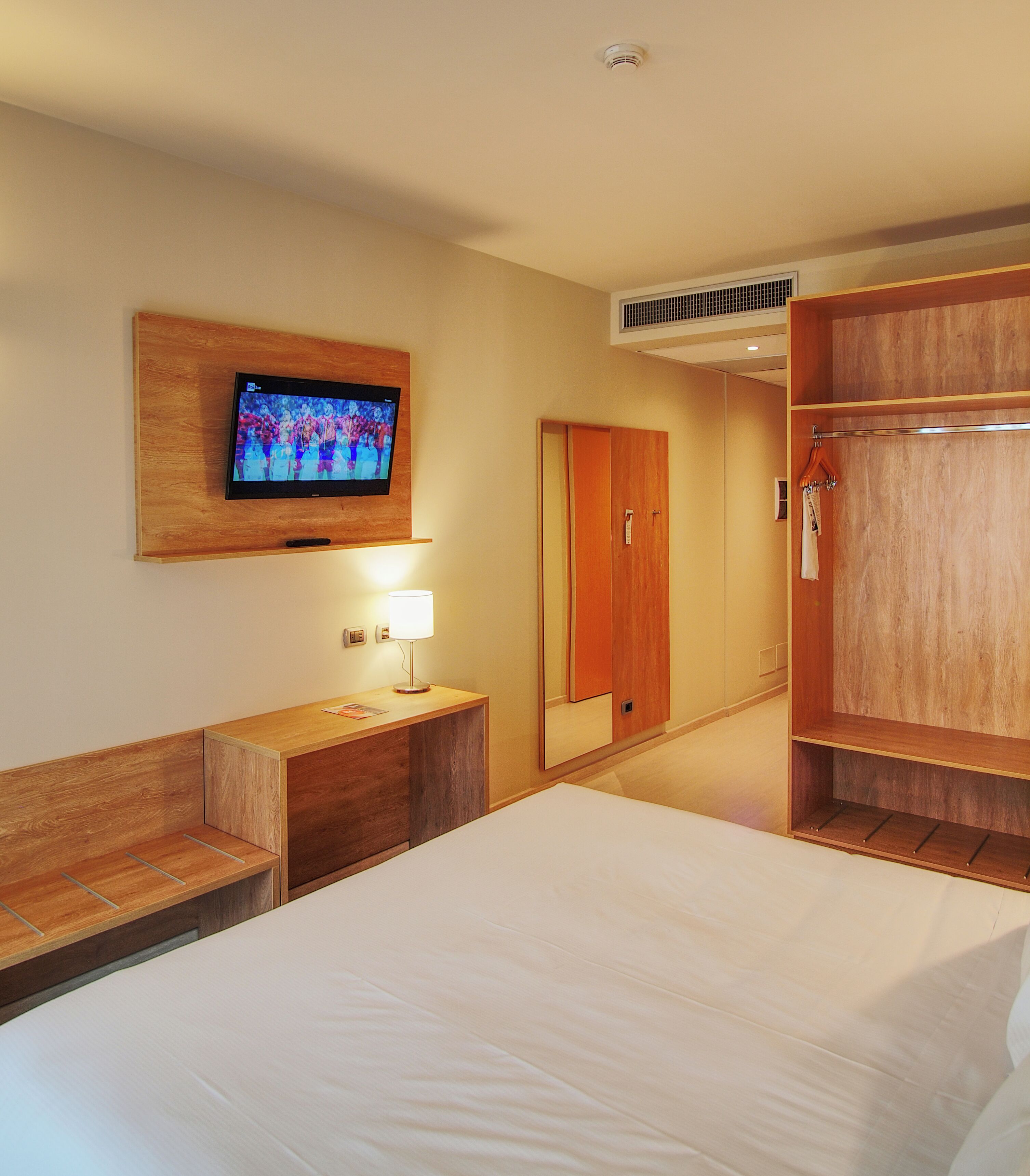Habitación de hotel con cama, televisión montada en la pared, mesita de noche con lámpara, armario y espejo.