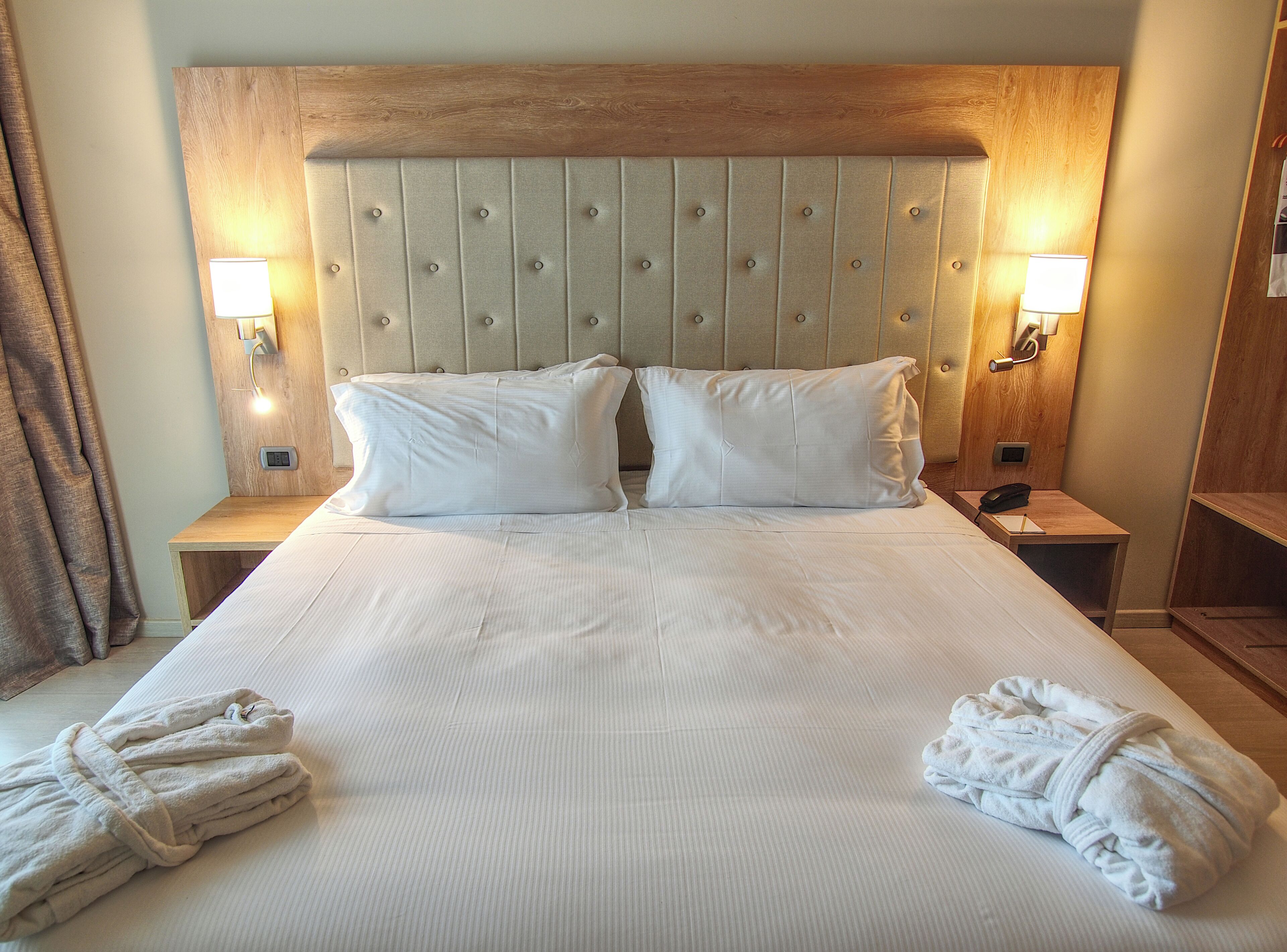 A hotel room with a large bed, two pillows, two bedside lamps, a wooden headboard, and two towels on the bed.